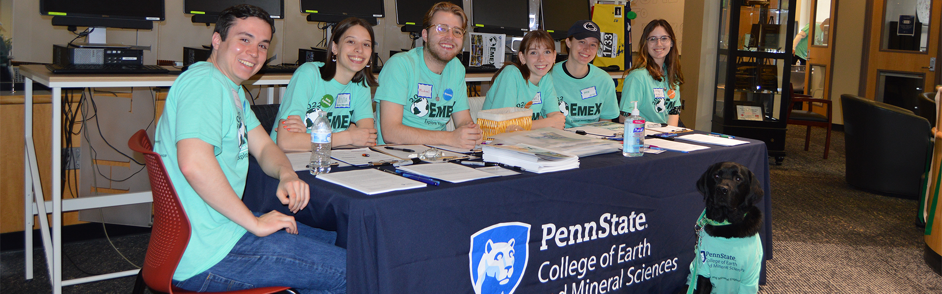 5 students and a dog sitting at a table and smiling and wearing EMEX 22 tshirts